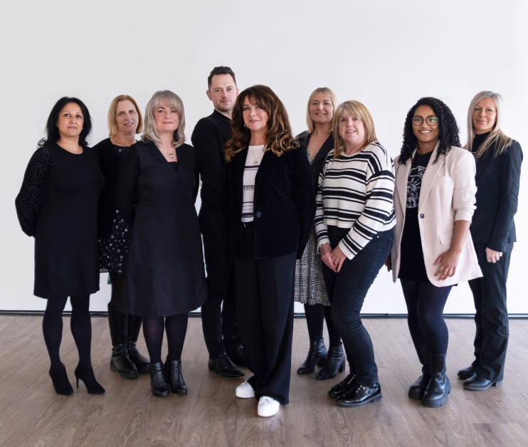 A group of eleven individuals stands together in a well-lit space with a plain white backdrop. They are dressed in a mix of formal and casual clothing, with varying styles and colors, including black, white, and patterned garments. The group displays a range of hairstyles and poses, looking towards the camera.