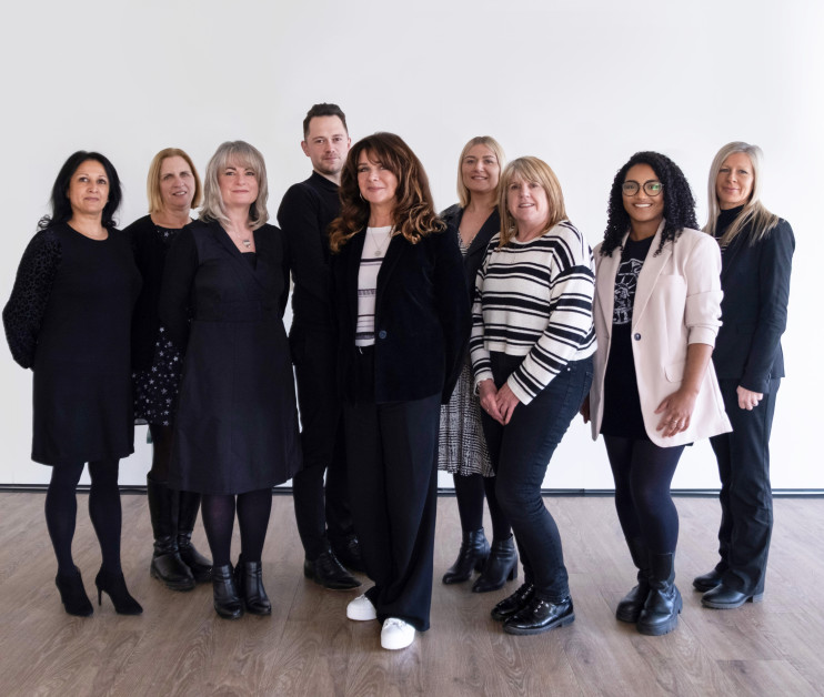 A group of ten individuals stands together in a bright, minimalist setting. They are dressed in a variety of styles, predominantly in dark and neutral colors, with some wearing patterns. The group appears to be smiling and engaged, showcasing a diverse mix of ages and hairstyles.