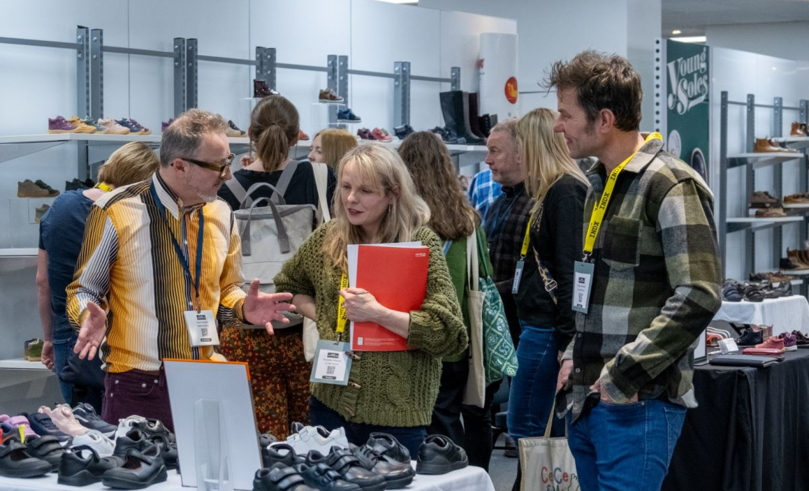 A group of three people engaged in conversation at a shoe exhibition. One man is gesturing while discussing, a woman is holding a red folder and looking intently, and another man listens while standing next to her. Various shoe displays are visible in the background.