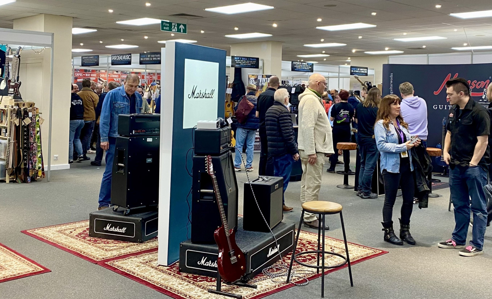 A bustling music exhibition featuring various attendees exploring booths. Prominent Marshall amplifiers and guitar displays are set up in the foreground, with people engaged in conversation and browsing merchandise in the background.