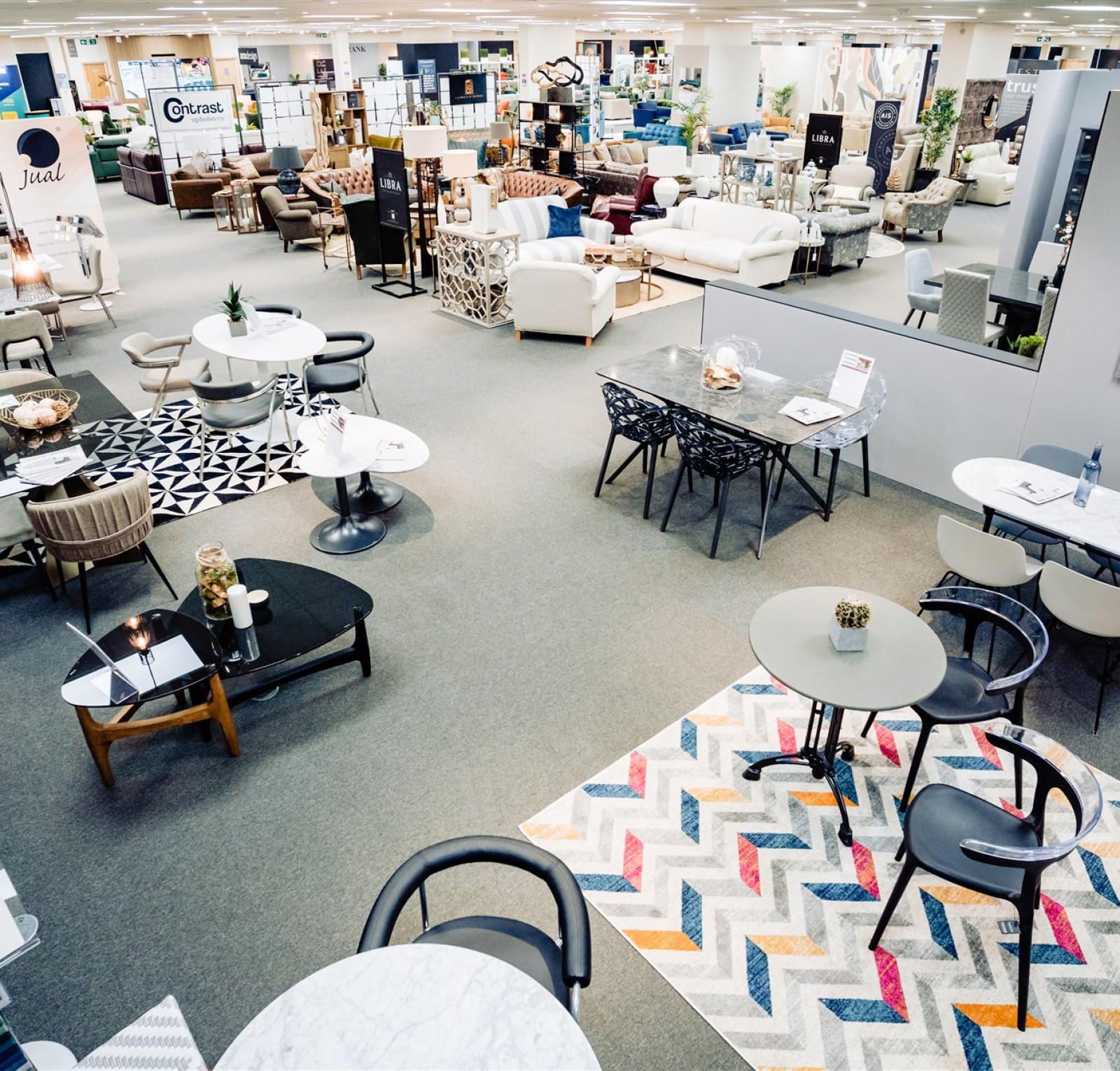 A modern furniture store interior showcases various seating arrangements and tables. Different styles of rugs add color to the space, while displays feature a mix of chairs and decorative items. The open layout highlights a range of furniture pieces for living and dining areas.