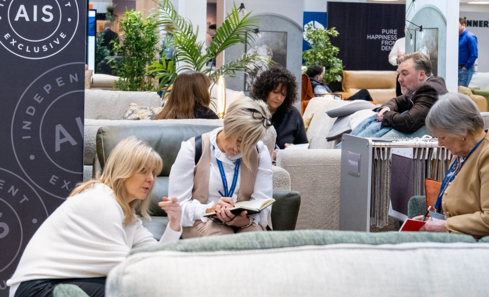 A group of people seated in a modern furniture showroom, engaged in discussions and taking notes. Several stylish sofas and plants are visible in the background. The environment appears relaxed and collaborative.
