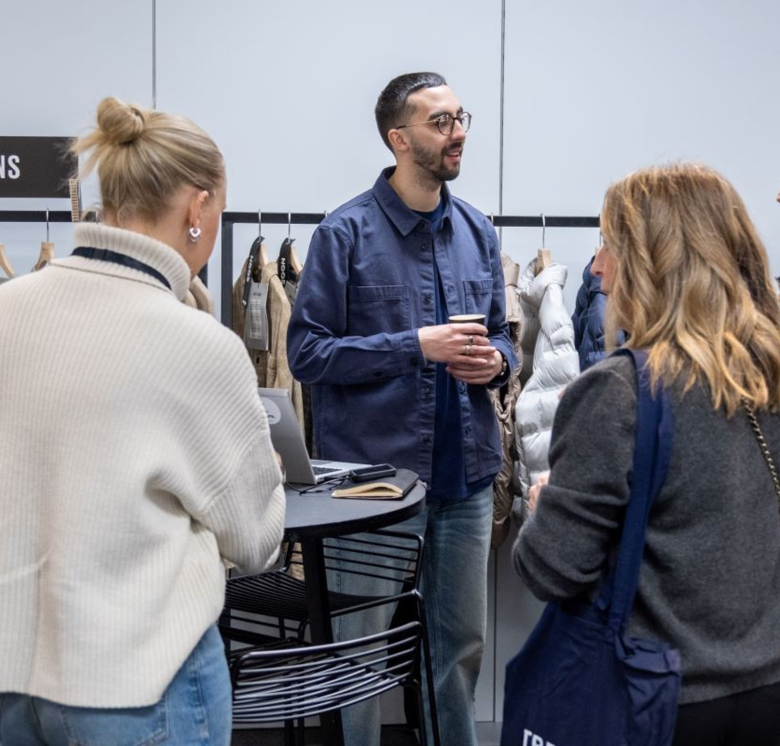 A man in glasses and a blue shirt engages with a small group of people at a clothing display. They are talking near a rack of hanging garments, with a table and laptop in front of them.