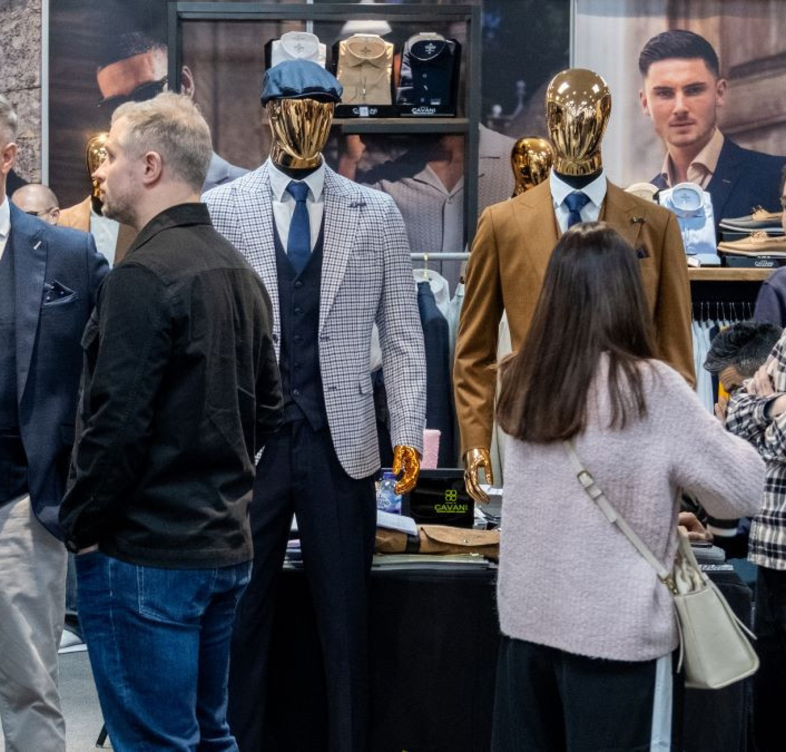 A crowd of people at a fashion exhibition, featuring well-dressed mannequins in stylish suits. Attendees are engaged in conversation, surrounded by fashion displays and promotional materials.