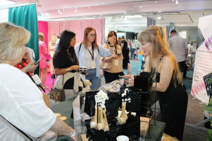 A woman demonstrates products at a vendor booth while a group of interested customers, including women and a man, look on. The booth features various beauty tools and accessories displayed on a table. Colorful backdrops and banners are visible in the background.