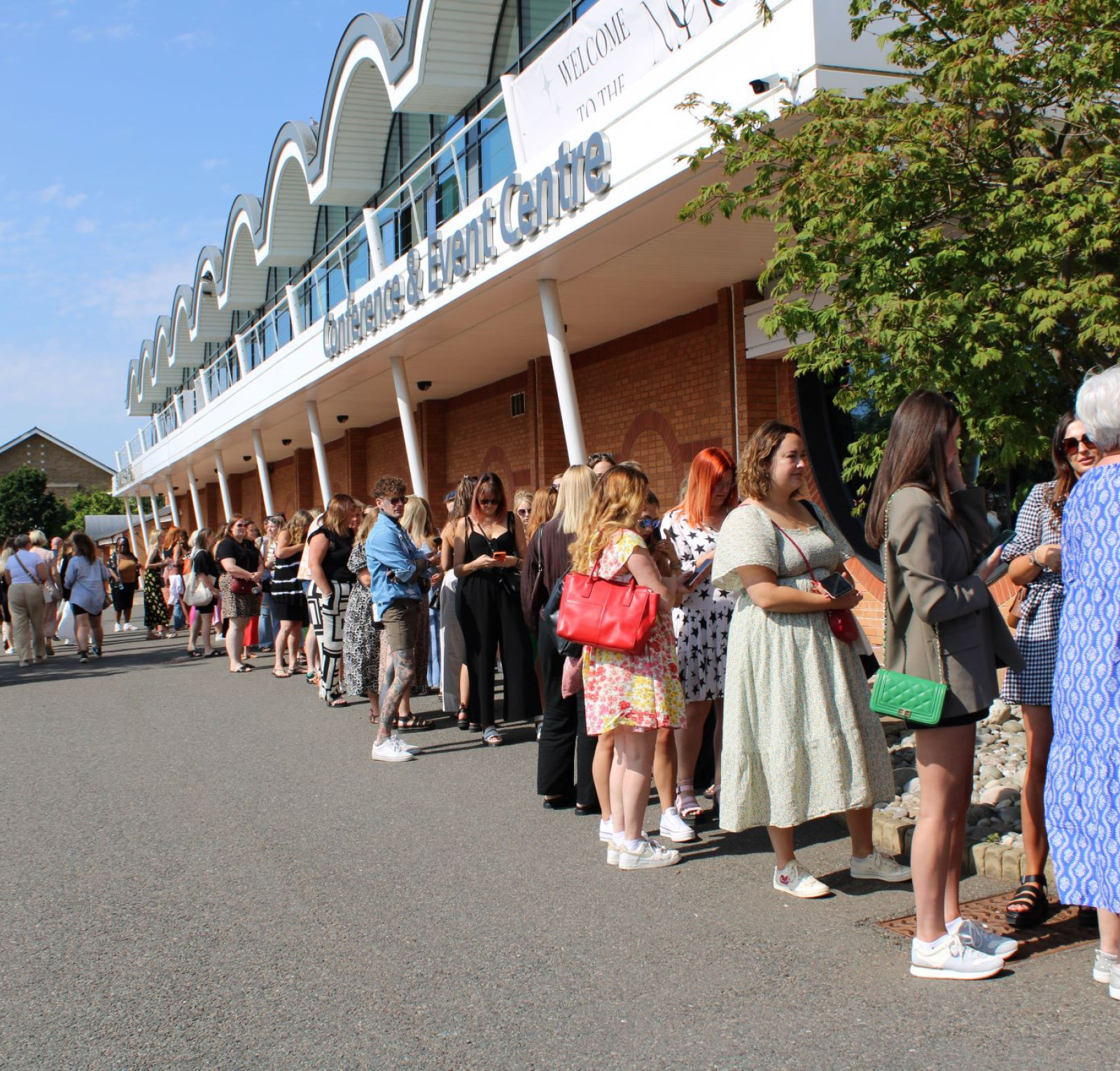 A long line of people waiting outside a building on a sunny day. The crowd is diverse, with individuals wearing various outfits, including dresses and casual attire. Trees and parked cars are visible along the pathway.