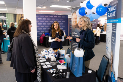 A booth at a health fair features a representative speaking with a visitor. The booth displays eye care products and promotional materials, with blue balloons in the background. Two women are engaged in conversation, while a table showcases various eye care items and brochures.
