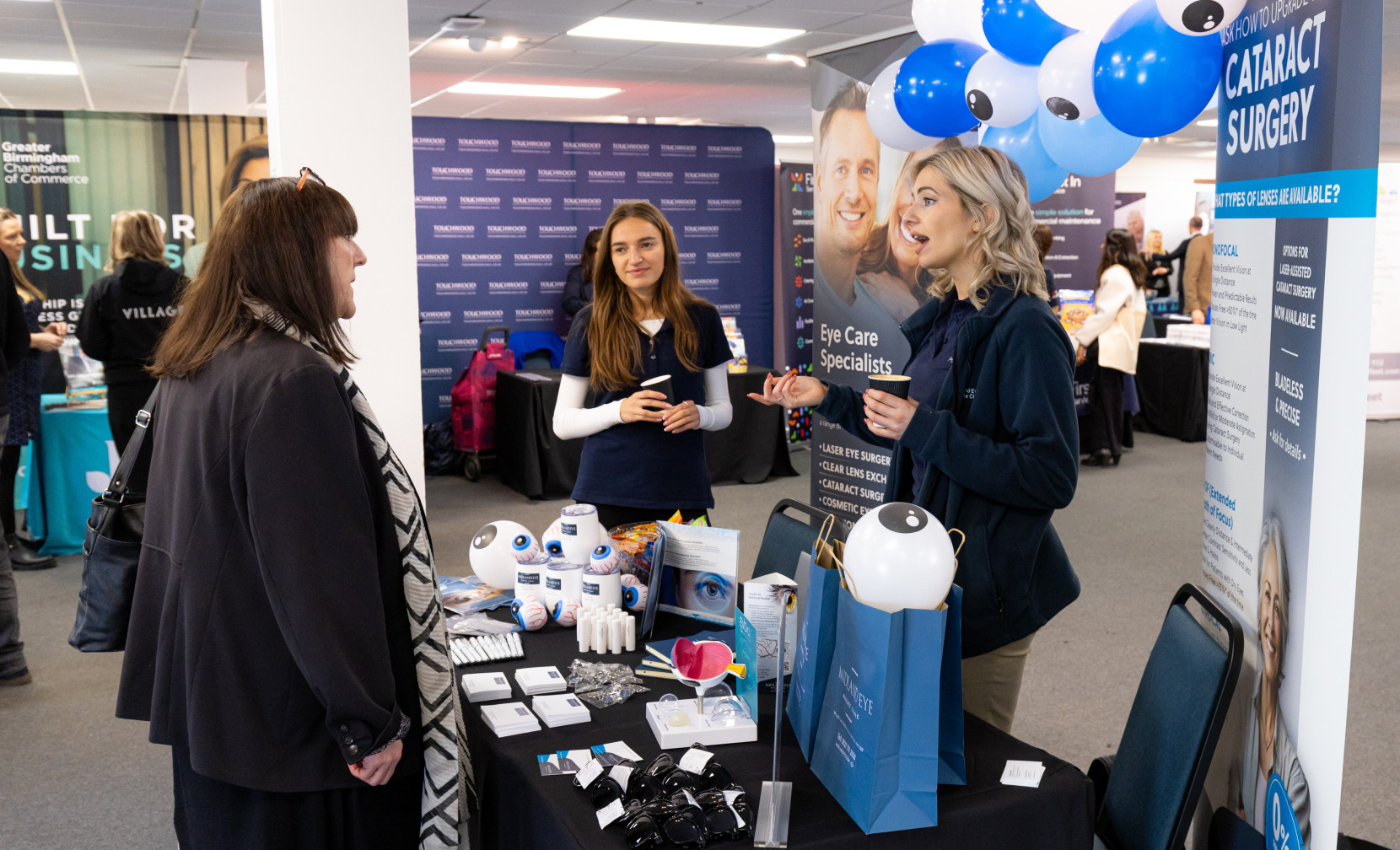 A booth at a health fair features a representative speaking with a visitor. The booth displays eye care products and promotional materials, with blue balloons in the background. Two women are engaged in conversation, while a table showcases various eye care items and brochures.