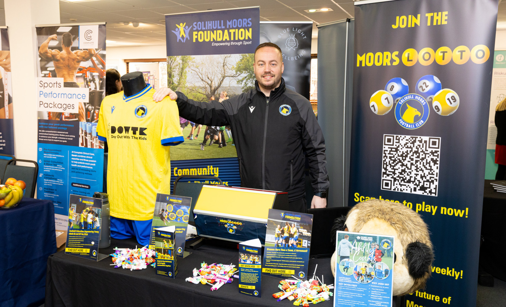 A man stands next to a display table featuring a yellow soccer jersey with blue accents, promotional materials for the Solihull Moors Foundation, and a mascot. The backdrop includes a large banner with a QR code and information about the Moors Lottery.