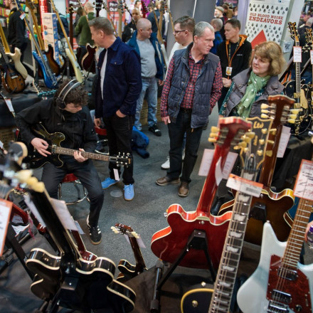 A crowded guitar exhibition with various electric and acoustic guitars on display. A person plays an electric guitar while others observe and browse the instruments. The setting shows attendees engaged in the music event.