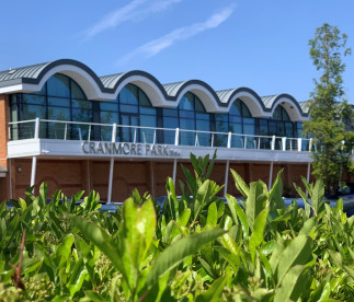 Cranmore Park building with a distinctive wavy roof design, featuring large glass windows and a balcony, surrounded by lush green foliage under a clear blue sky.
