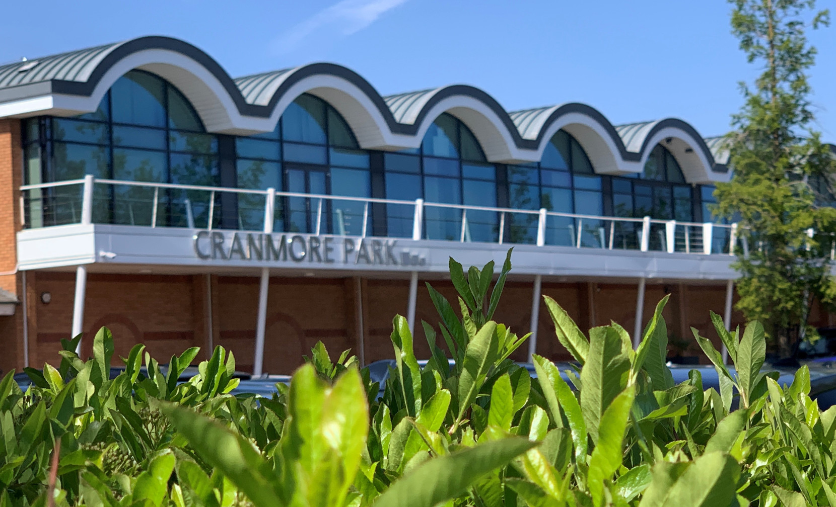 Cranmore Park building with a distinctive wavy roof design, featuring large glass windows and a balcony, surrounded by lush green foliage under a clear blue sky.