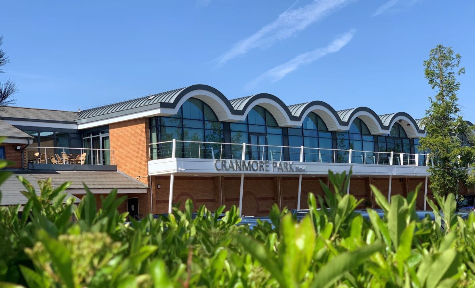 Cranmore Park building with a curved roof design, featuring large windows and a balcony. Clear blue sky and greenery in the foreground.