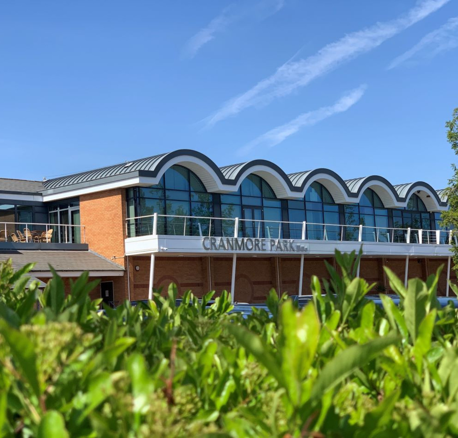 Cranmore Park building with a curved roof design, featuring large windows and a balcony. Clear blue sky and greenery in the foreground.