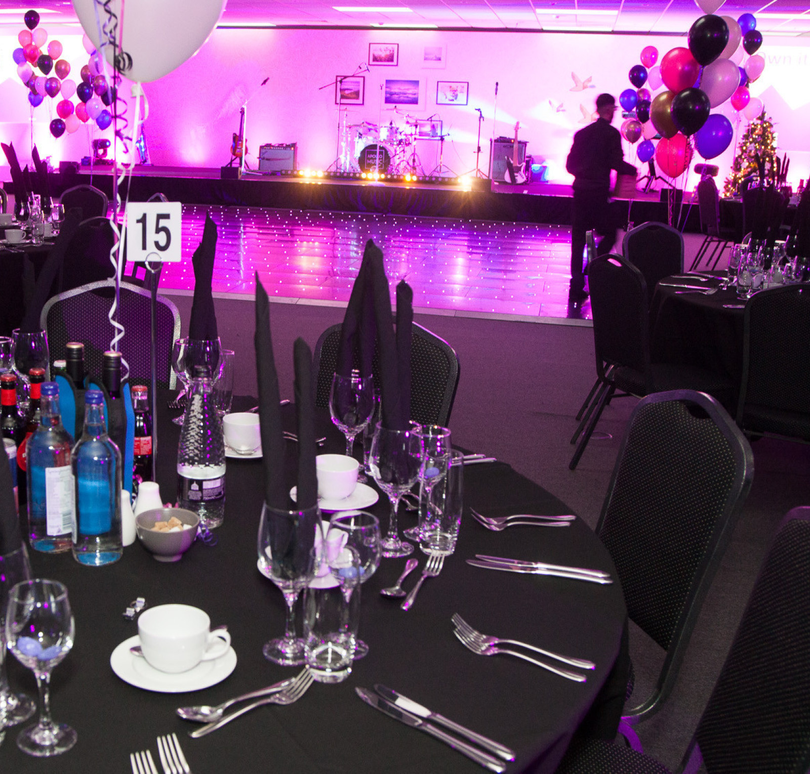 A formal dining setup featuring tables with black tablecloths, neatly arranged cutlery, and drinks. In the background, a stage with musical instruments is illuminated by colorful lights, and balloons in various colors are displayed around the room.