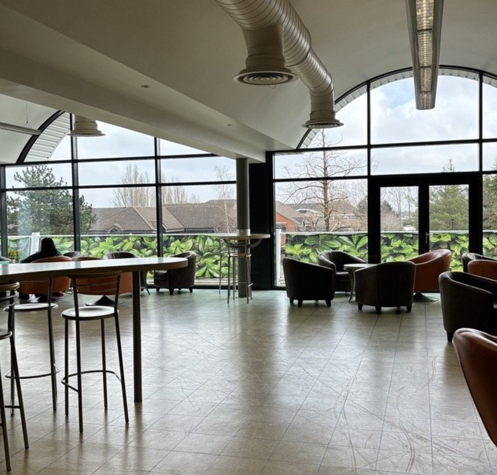 A spacious indoor seating area features modern furniture, including brown armchairs and high-top tables. Large windows provide natural light and a view of greenery outside. Soft, neutral-colored walls and a contemporary ceiling design create a welcoming atmosphere.