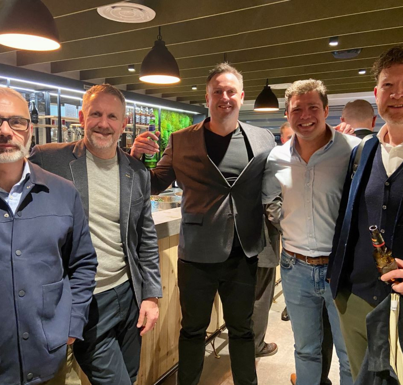 A group of five men stands together in a stylish bar. They are smiling and dressed in smart casual attire. The background features shelves filled with various drinks, and the ambiance is warm with pendant lighting.