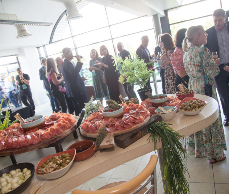 A vibrant gathering takes place in a stylish venue, featuring guests mingling and enjoying drinks. In the foreground, a beautifully arranged spread of charcuterie, including cured meats, olives, and garnishes, invites appetites. Natural light streams in through large windows, enhancing the lively atmosphere.