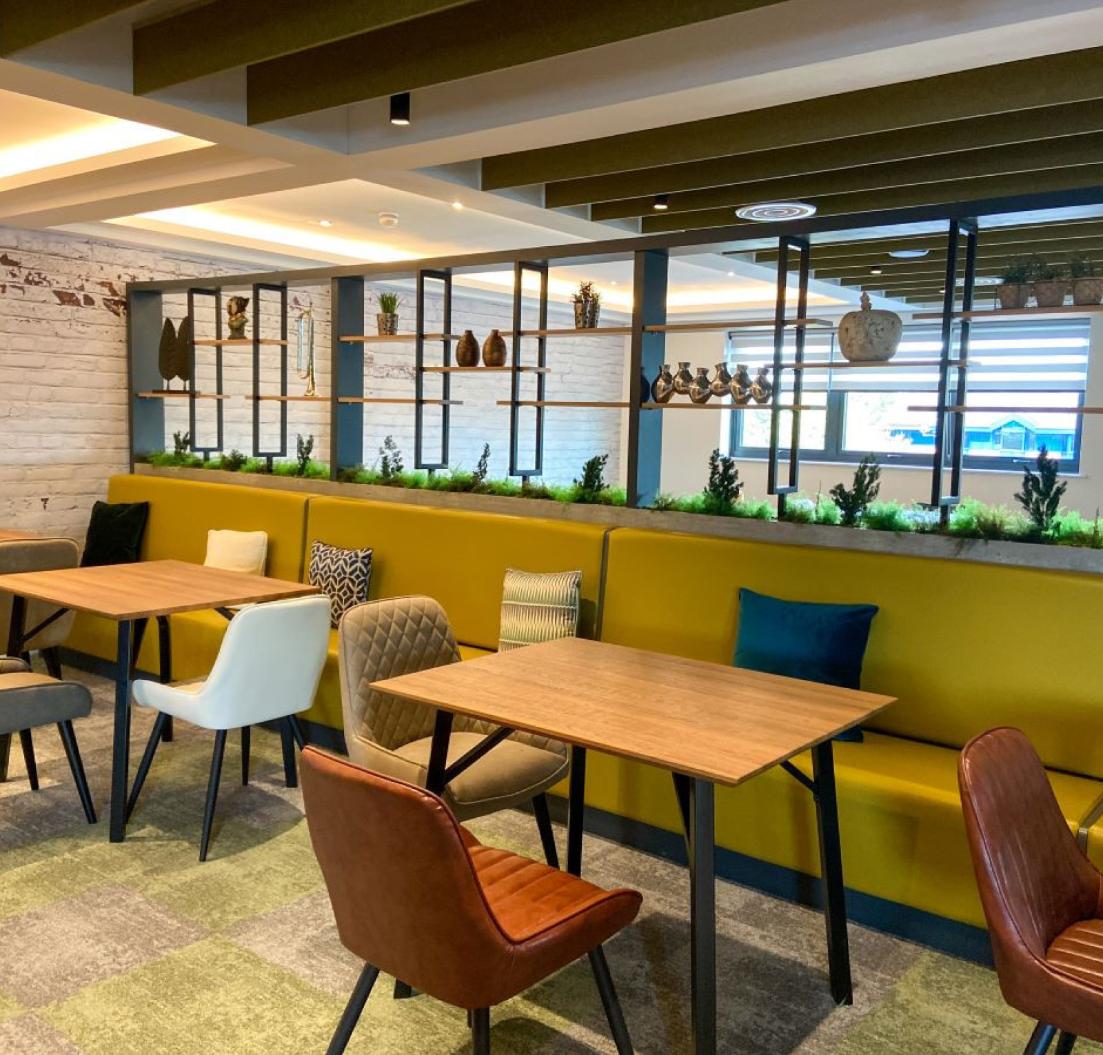 A modern cafe interior featuring wooden tables and a variety of seating options, including upholstered chairs. Greenery accents the space, with shelves displaying decorative items in the background. Soft lighting enhances the inviting atmosphere.