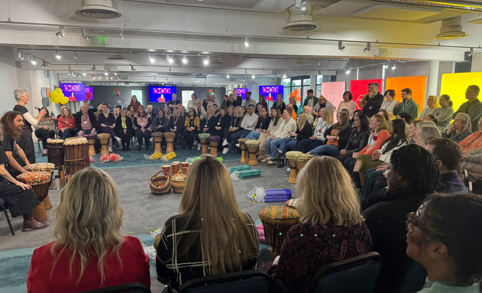 A large group of people seated in a circular arrangement, engaged in a drumming activity led by two facilitators. Colorful decorations and screens are visible in the background. People of various ages and ethnicities interact with drums and each other in a collaborative environment.