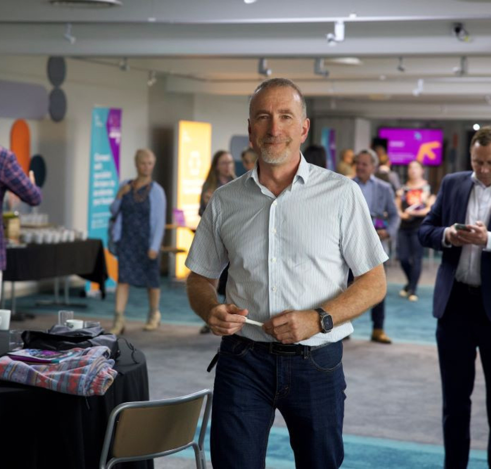 A man in a light blue shirt stands confidently in a modern event space, smiling at the camera. In the background, attendees engage in conversation and coffee service is visible. Bright banners decorate the room, adding to the lively atmosphere.