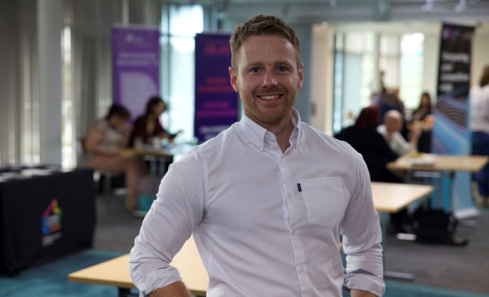 A smiling man in a white shirt stands confidently in a well-lit modern workspace. Background features blurred tables and colorful banners, with people engaged in conversation.