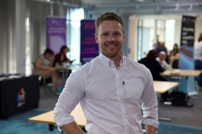 A smiling man in a white shirt stands confidently in a well-lit modern workspace. Background features blurred tables and colorful banners, with people engaged in conversation.