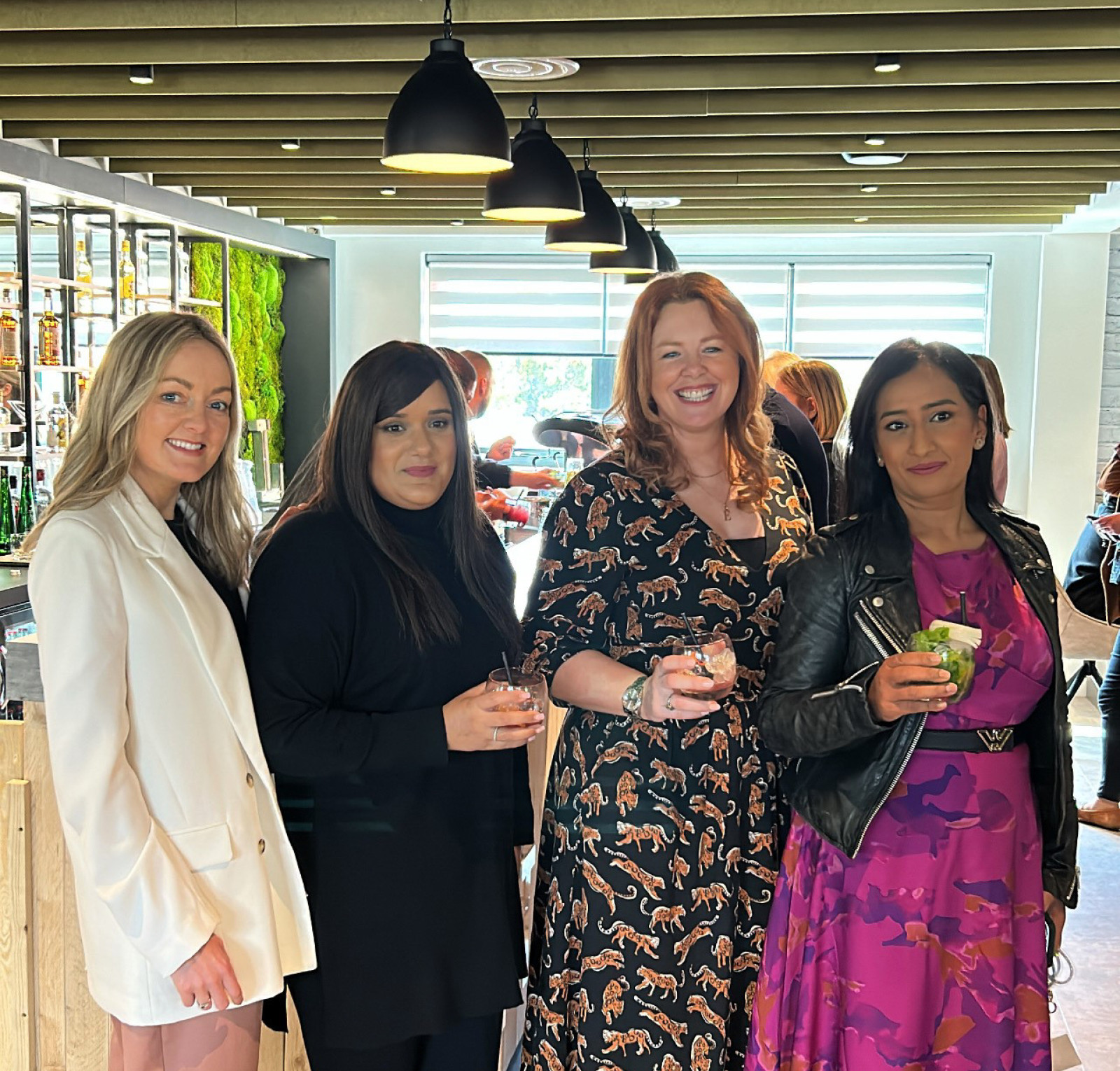 Four women stand together in a modern setting, each holding a drink. They are dressed in stylish outfits, with two in dresses and the others in a blazer and casual wear. The background features a bar area with greenery.