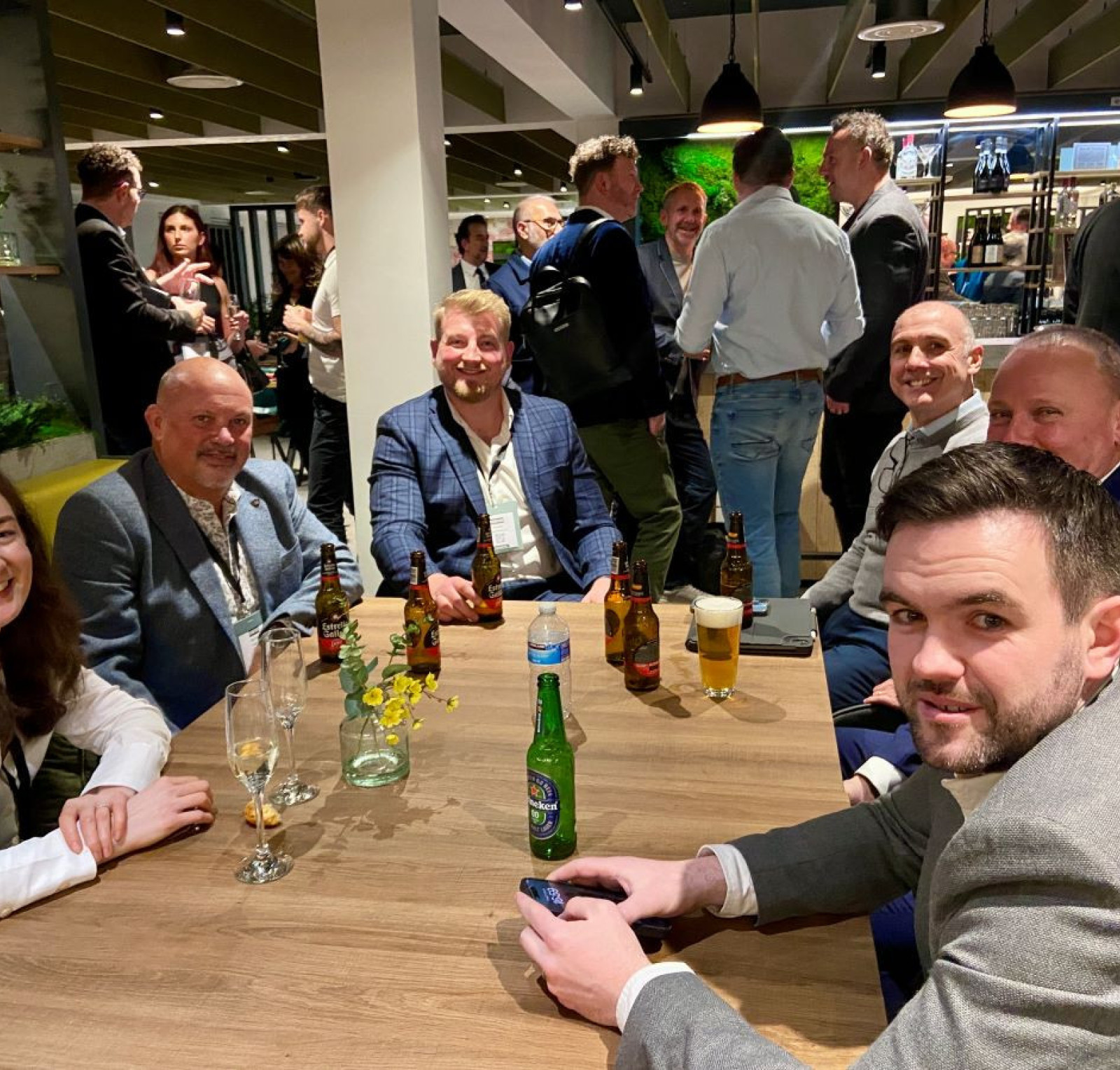 A group of six people sitting around a wooden table in a lively bar atmosphere. They are enjoying drinks and engaging in conversation, with a mixture of casual and more formal attire. In the background, other patrons are socializing.