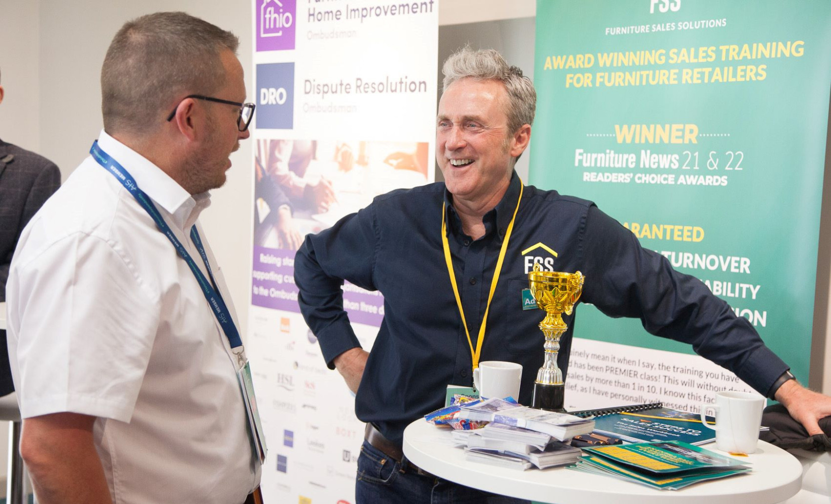Two men engage in conversation at a trade event. One man holds a trophy and smiles, wearing a navy shirt with a logo, while the other looks on attentively in a white shirt and glasses. A banner in the background highlights awards and sales training for furniture retailers.
