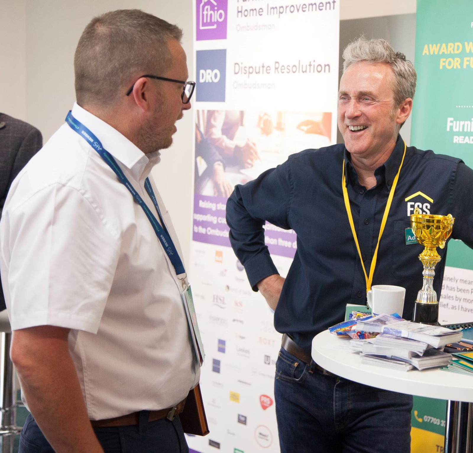 Two men engage in conversation at a trade event. One man holds a trophy and smiles, wearing a navy shirt with a logo, while the other looks on attentively in a white shirt and glasses. A banner in the background highlights awards and sales training for furniture retailers.