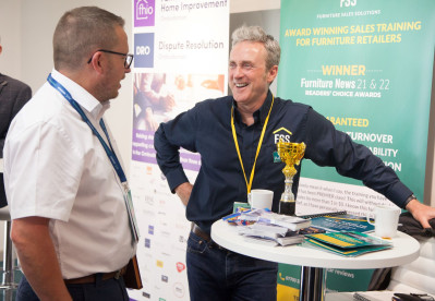 Two men engage in conversation at a trade event. One man holds a trophy and smiles, wearing a navy shirt with a logo, while the other looks on attentively in a white shirt and glasses. A banner in the background highlights awards and sales training for furniture retailers.