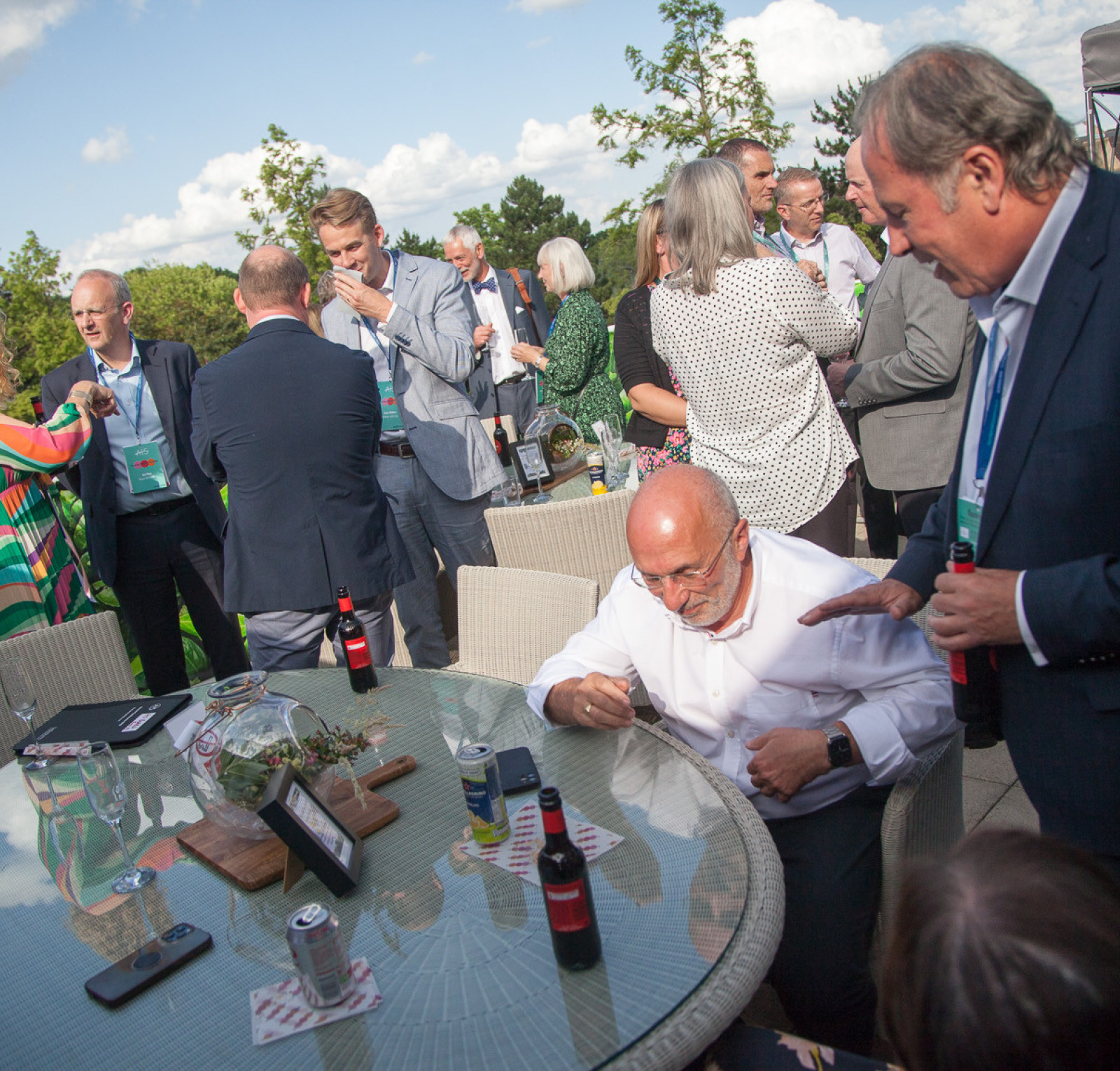 A lively outdoor gathering with people mingling around a glass table. Some individuals are seated, while others stand in conversation, enjoying refreshments. The background features green trees and a partly cloudy sky, creating a relaxed atmosphere.