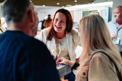 A woman in a white blazer smiles while holding a glass of champagne, engaged in conversation with two people at a social event. Bright, modern decor is visible in the background, indicating a lively atmosphere.