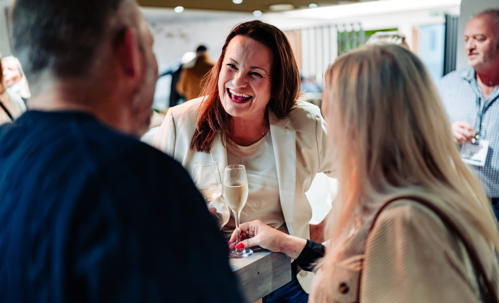 A woman in a white blazer smiles while holding a glass of champagne, engaged in conversation with two people at a social event. Bright, modern decor is visible in the background, indicating a lively atmosphere.