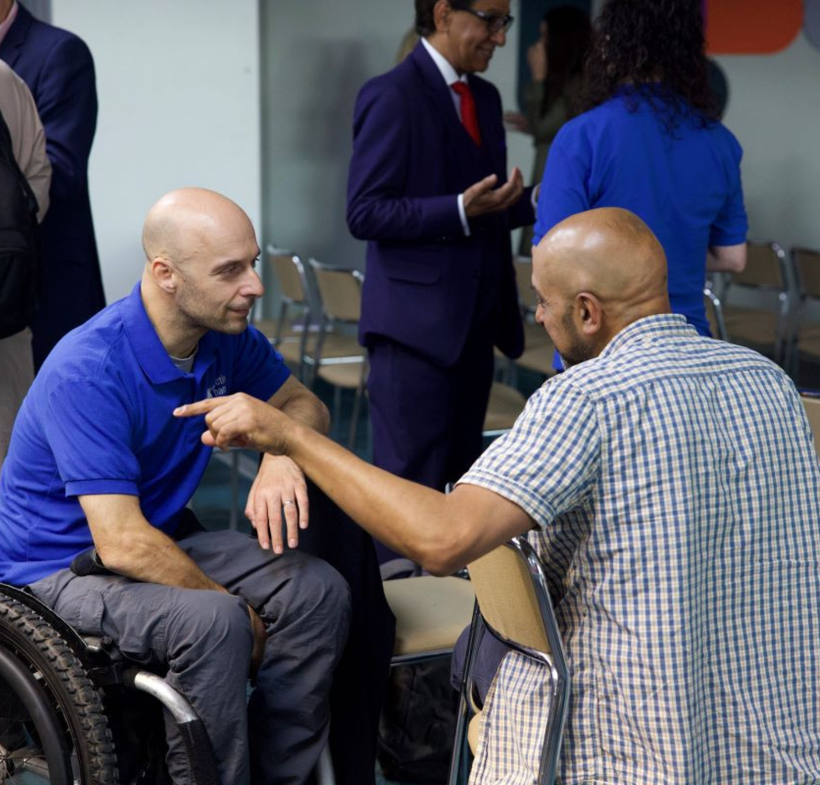 Two men engage in a discussion in a conference setting. One man, seated in a wheelchair, wears a blue shirt and looks attentively at the other man, who is standing and wearing a checkered shirt. In the background, a few other individuals are mingling.