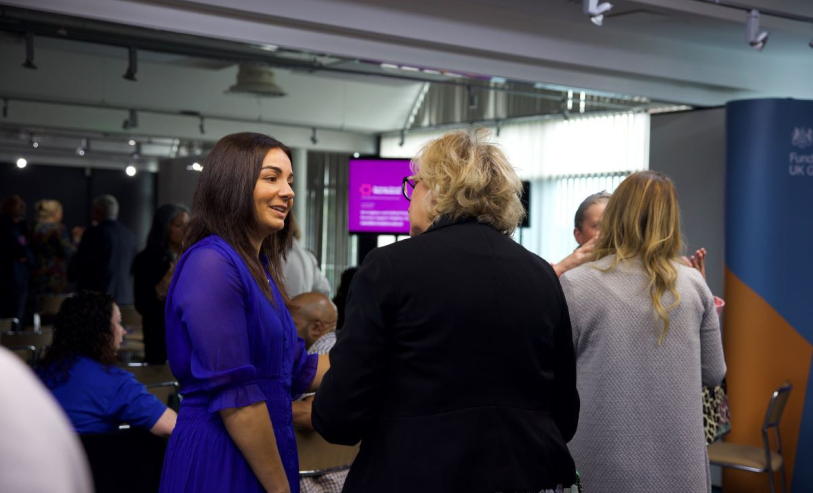 Two women engage in conversation at an indoor event, with a group of people mingling in the background. One woman is dressed in a blue dress, while the other wears a black jacket. A screen displaying information can be seen in the distance.