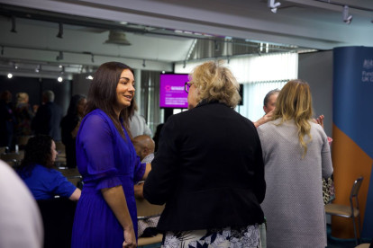 Two women engage in conversation at an indoor event, with a group of people mingling in the background. One woman is dressed in a blue dress, while the other wears a black jacket. A screen displaying information can be seen in the distance.