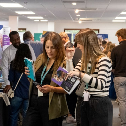 A busy event space with various vendors and attendees engaged in conversations. Two women in the foreground are examining brochures and materials, while people browse booths in the background. Promotional banners and tables are visible around the room.