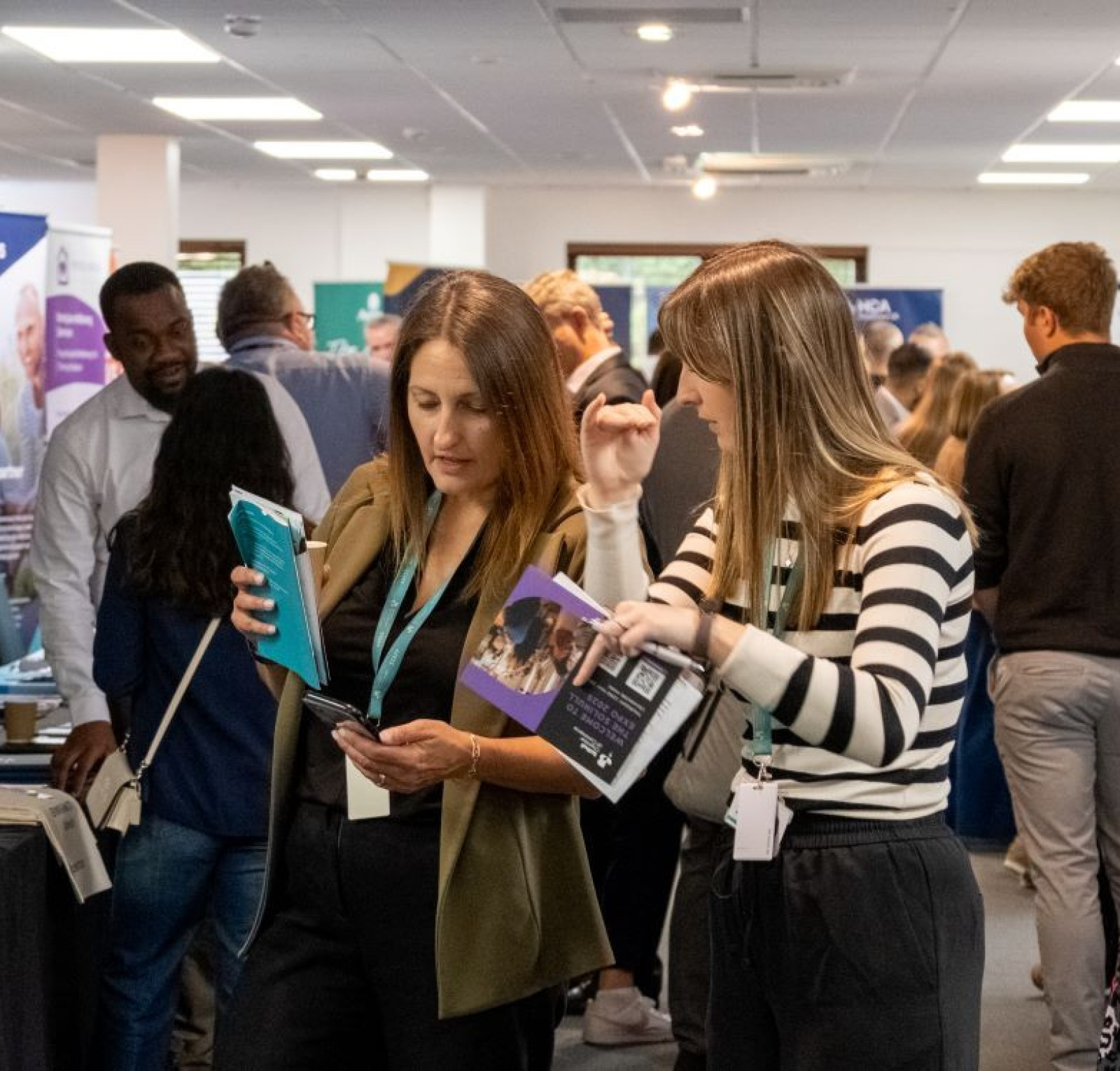 A busy event space with various vendors and attendees engaged in conversations. Two women in the foreground are examining brochures and materials, while people browse booths in the background. Promotional banners and tables are visible around the room.