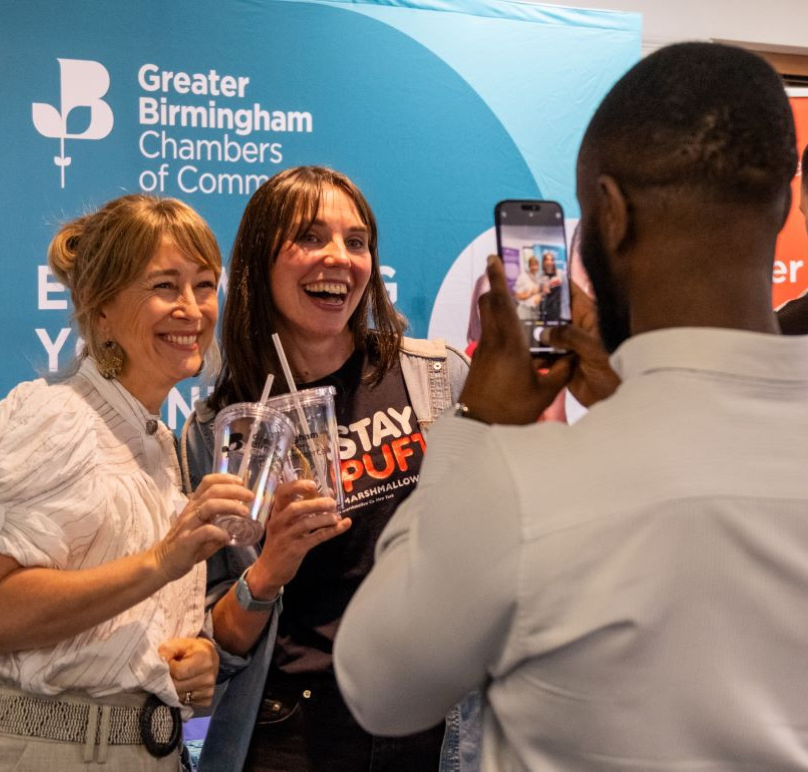Two women smile and pose together, holding drinks, while a man takes their picture with a smartphone. They are standing in front of a backdrop that features the Greater Birmingham Chambers of Commerce logo.
