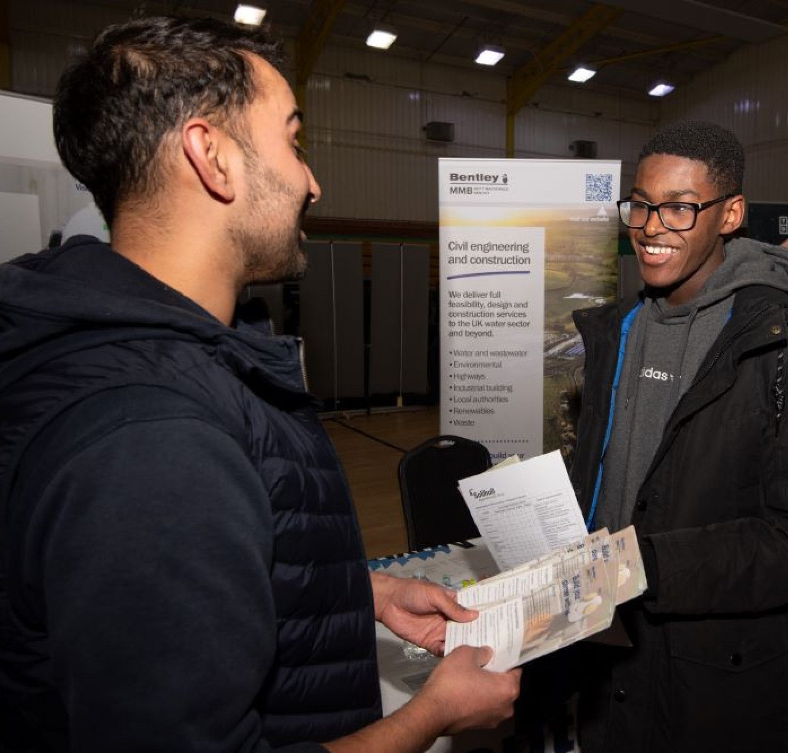 Two young men are engaged in conversation at a career fair. One man is handing a brochure to the other, who is smiling and listening attentively. Banners in the background showcase civil engineering and construction opportunities.