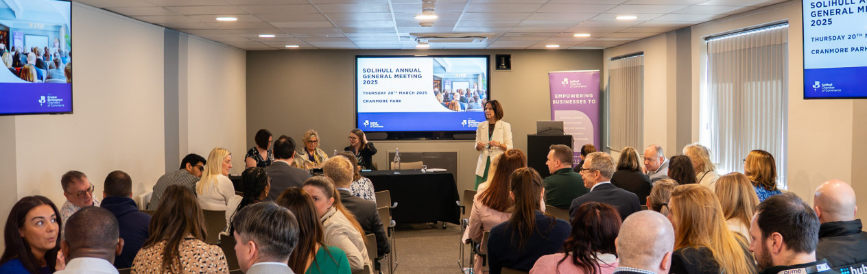 A conference room filled with attendees facing a presentation screen. A speaker stands at the front, engaging the audience during the annual general meeting. Informative visuals and banners are displayed around the room.