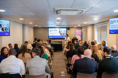 A conference room filled with attendees facing a presentation screen. A speaker stands at the front, engaging the audience during the annual general meeting. Informative visuals and banners are displayed around the room.