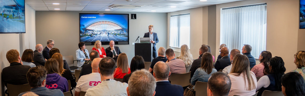 A speaker stands behind a podium in a conference room, addressing an audience seated in rows. The room features a large screen displaying a presentation, with several panelists seated to the left. Natural light streams in through windows.