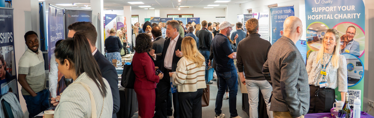 A busy event space filled with attendees networking and discussing at various booths. Tables display promotional materials, with individuals engaging in conversations throughout the area.