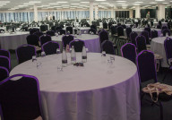A large conference hall set up with multiple round tables, each with water bottles and glasses. The space is brightly lit, featuring a backdrop of trees, and there are empty chairs around the tables.