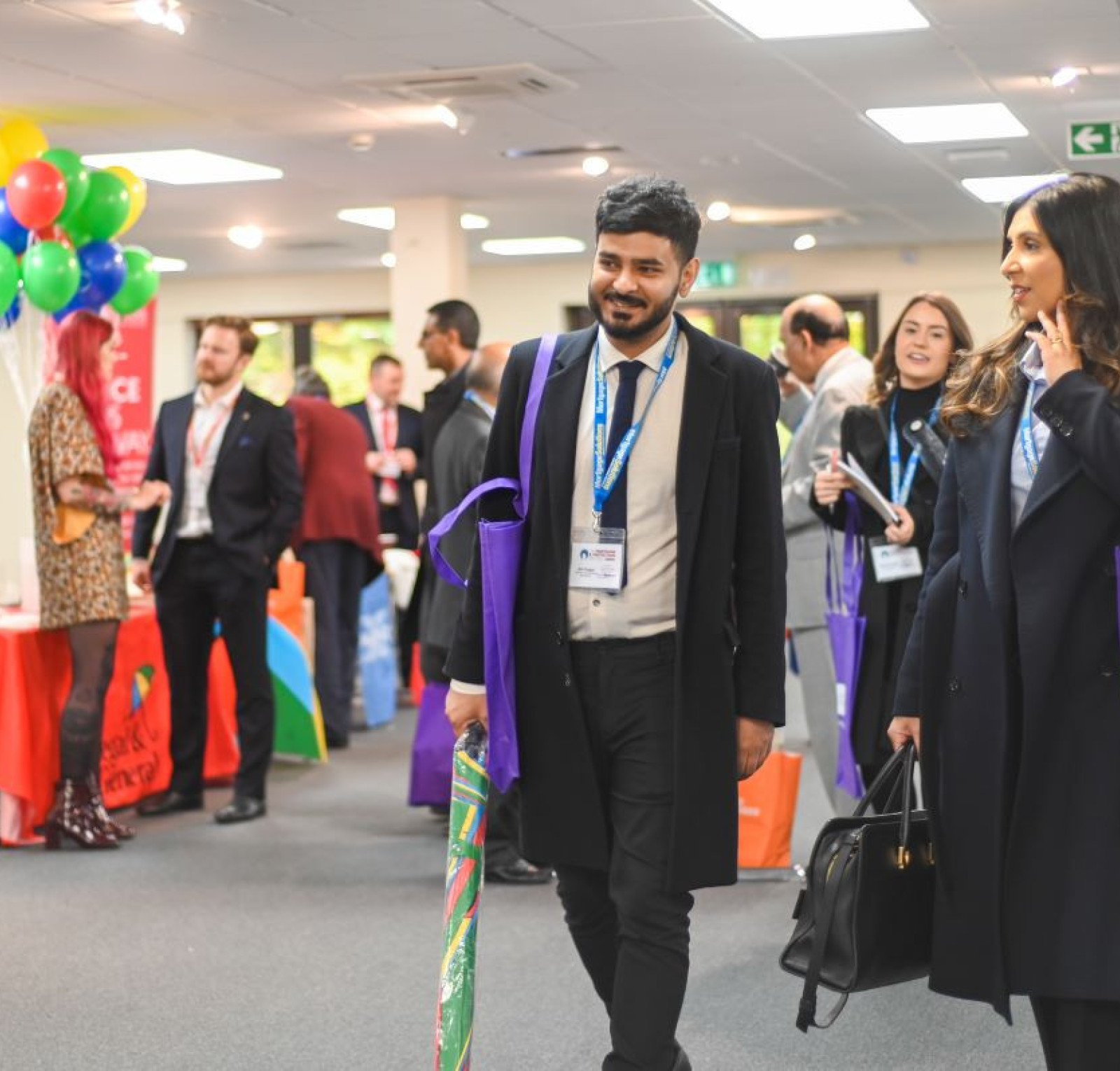 A group of professionals mingles at a networking event, with individuals carrying colorful bags. Balloons are in the background, and various exhibitor tables are set up around the venue.