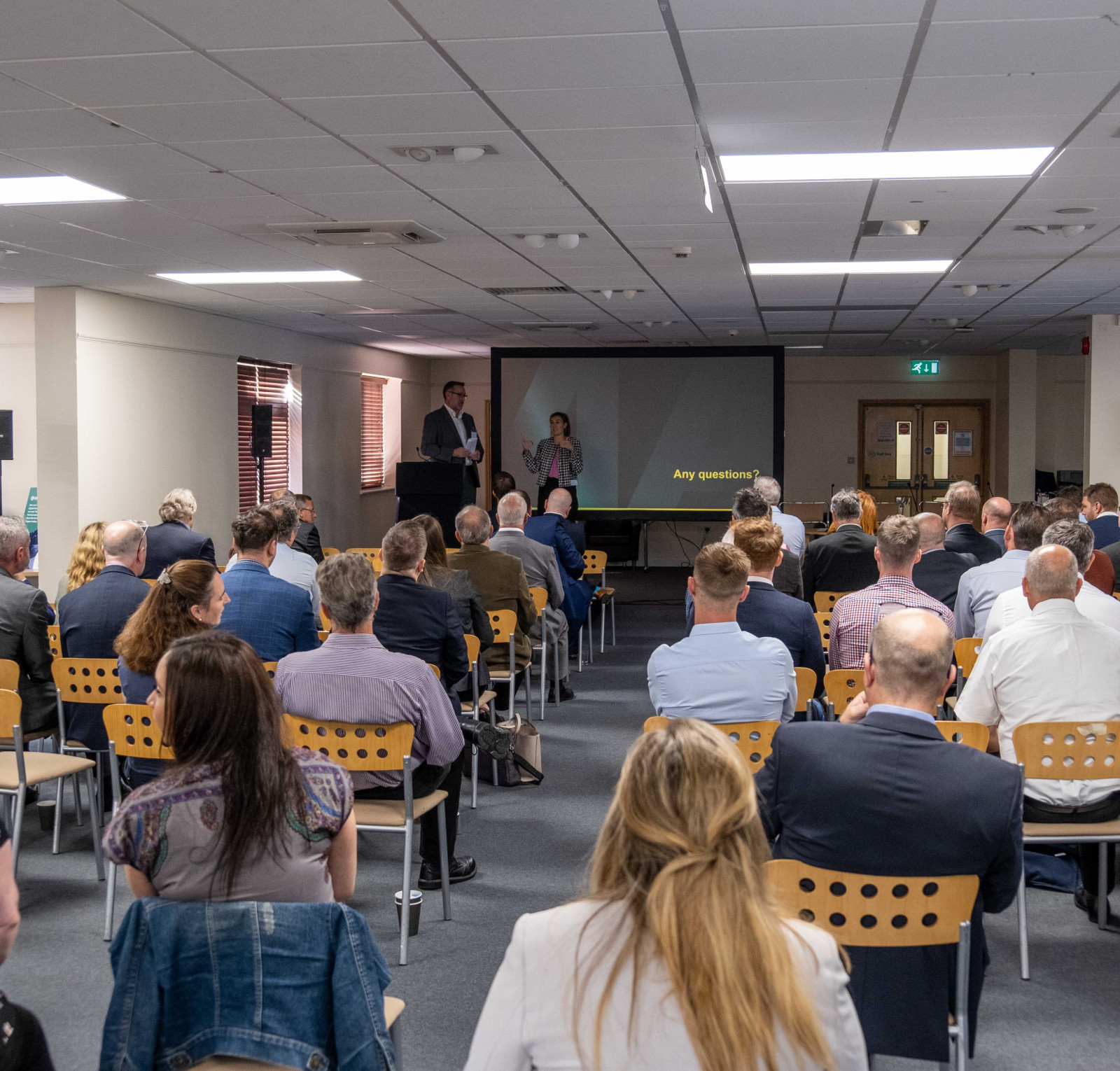 A crowded conference room with attendees seated, facing a presentation screen. Two speakers stand at the front, engaging the audience. Various people in business attire are positioned throughout the room, with a mix of men and women visible.
