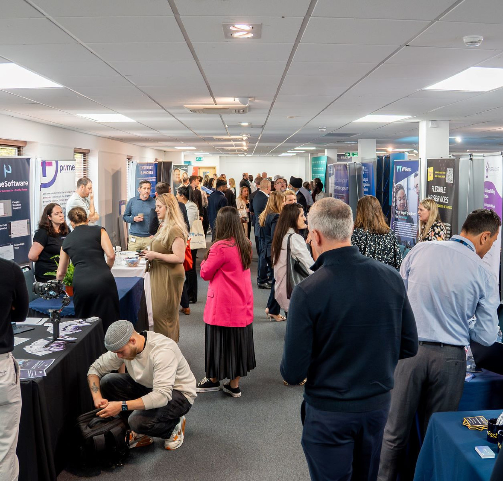 A bustling conference hall filled with people engaging at various booths. Attendees are networking and exploring displays about different programs and initiatives. Banners and signs promote services and skills training.